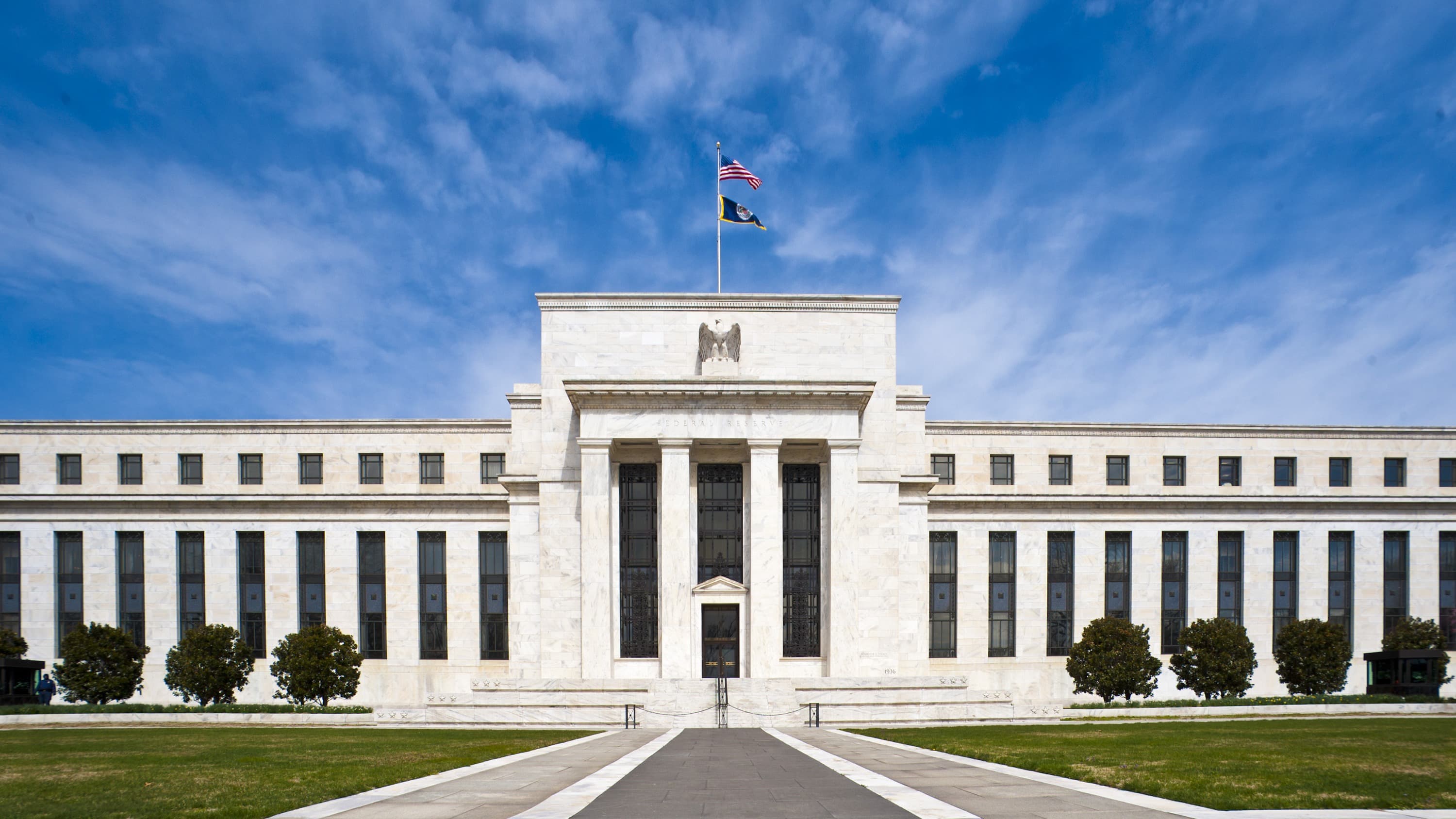 The white marble front of the Marriner S. Eccles Federal Reserve Board Building in Washington under a blue sky with thin clouds. A carved eagle sits above the central entrance, the words Federal Reserve are cut into the stone beneath it, and a United States flag and the Federal Reserve flag fly from a pole on the roof. A paved path leads across a lawn to the doors.