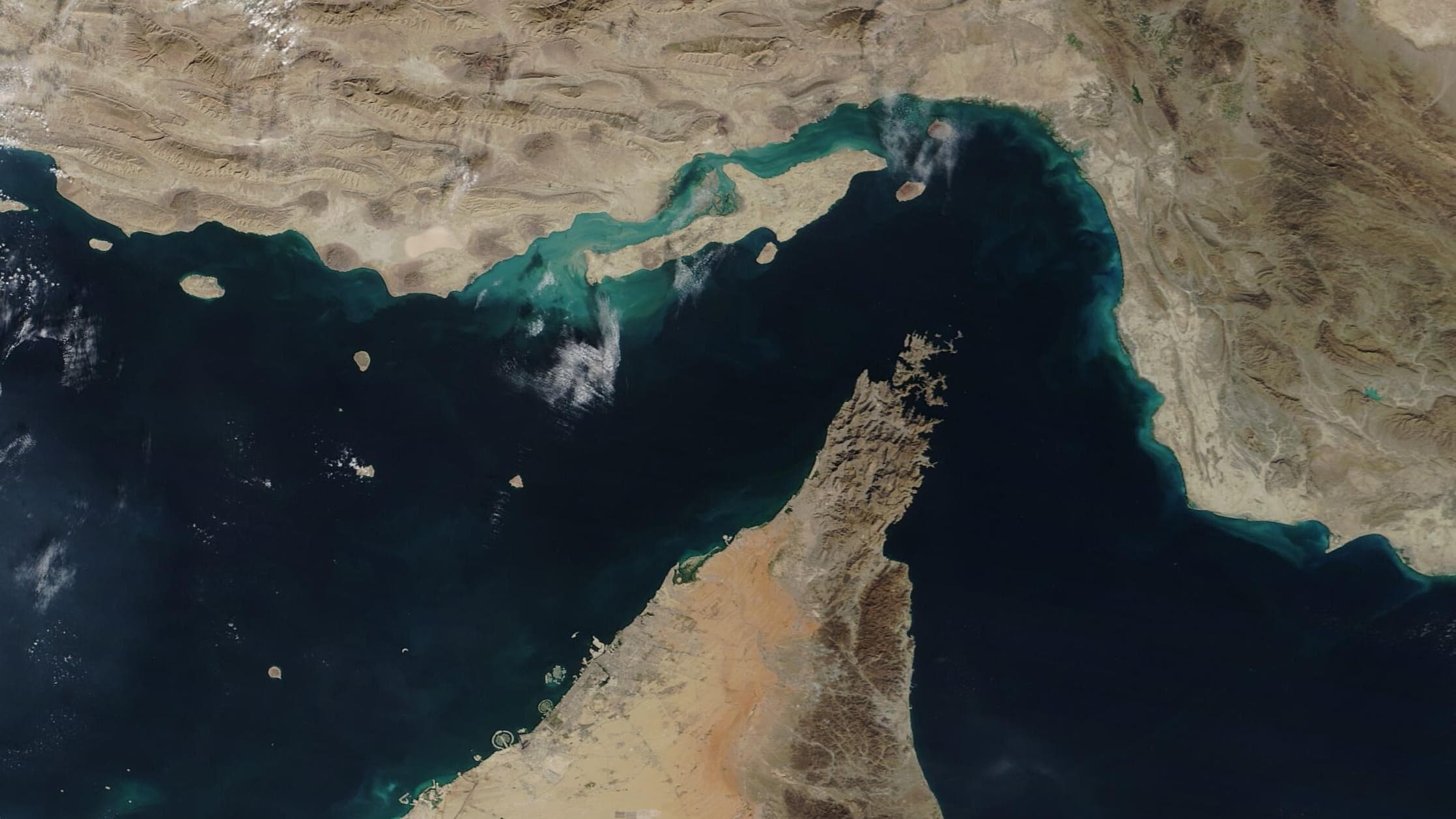 A satellite photograph of the Strait of Hormuz. Tan, ridged desert of the Iranian coast fills the top of the frame; below it the dark water of the Strait bends around the rocky Musandam Peninsula, which juts up from the bottom right; pale desert of the Arabian side sits at the bottom. Thin clouds drift over the water.