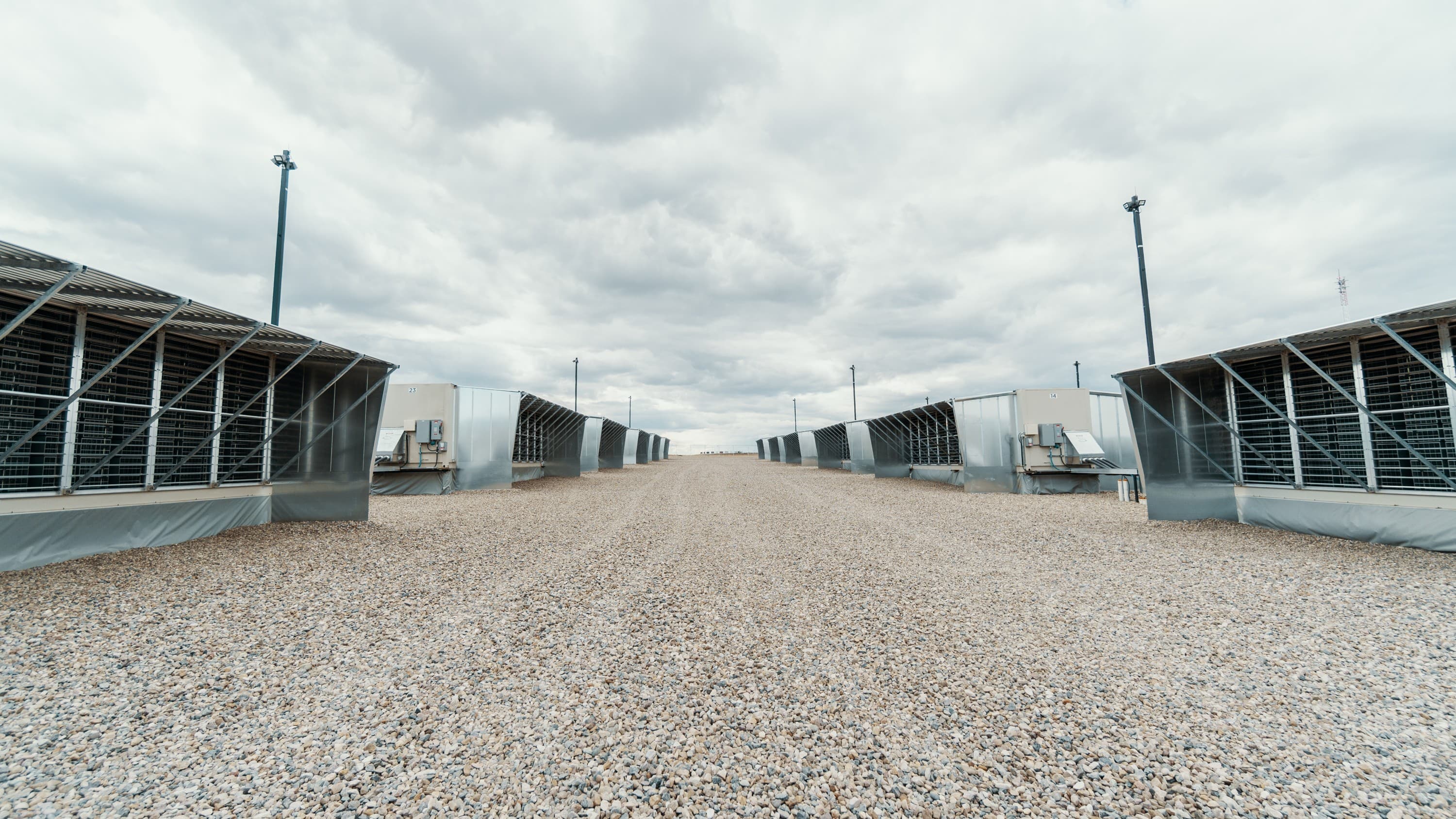 Two rows of steel shipping-container mining units with louvred sides face each other across a wide gravel yard, receding toward the horizon under a heavy overcast sky. Floodlight poles stand between them. The site is flat and empty of people.