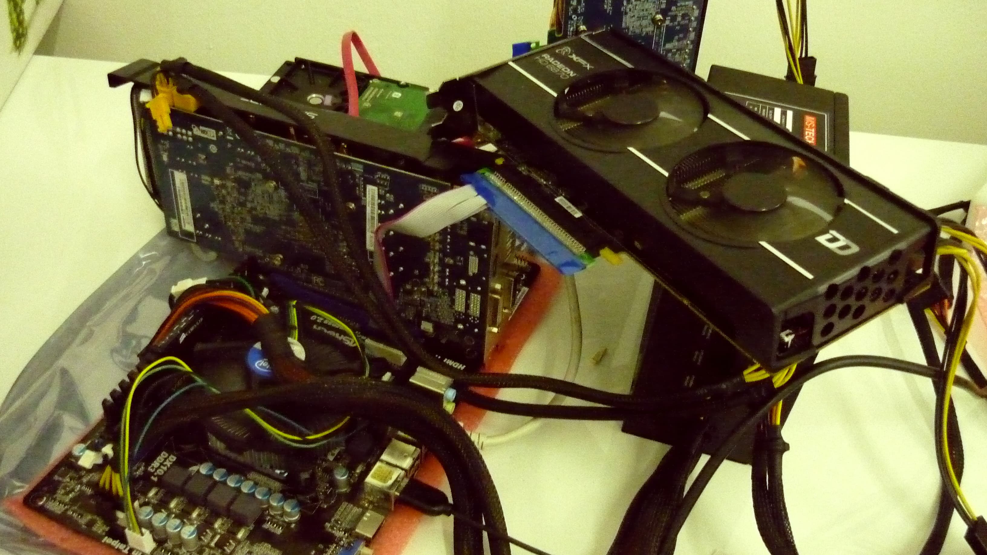 A home bitcoin mining rig on a white table, photographed in October 2012: two black Radeon graphics cards with twin fans stacked at angles over a bare motherboard with a round CPU cooler, a hard drive and a power supply, joined by braided and ribbon cables, with a pink anti-static mat under the board.